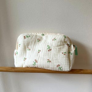 Könnte beinhalten: Eine weiße, rechteckige Kosmetiktasche mit floralem Muster. Die Tasche besteht aus einem strukturierten Stoff und weist kleine, gestickte rote und grüne Blumen auf. Sie hat einen beigen Reißverschluss und eine kleine grüne Stoffschlaufe an der Seite.