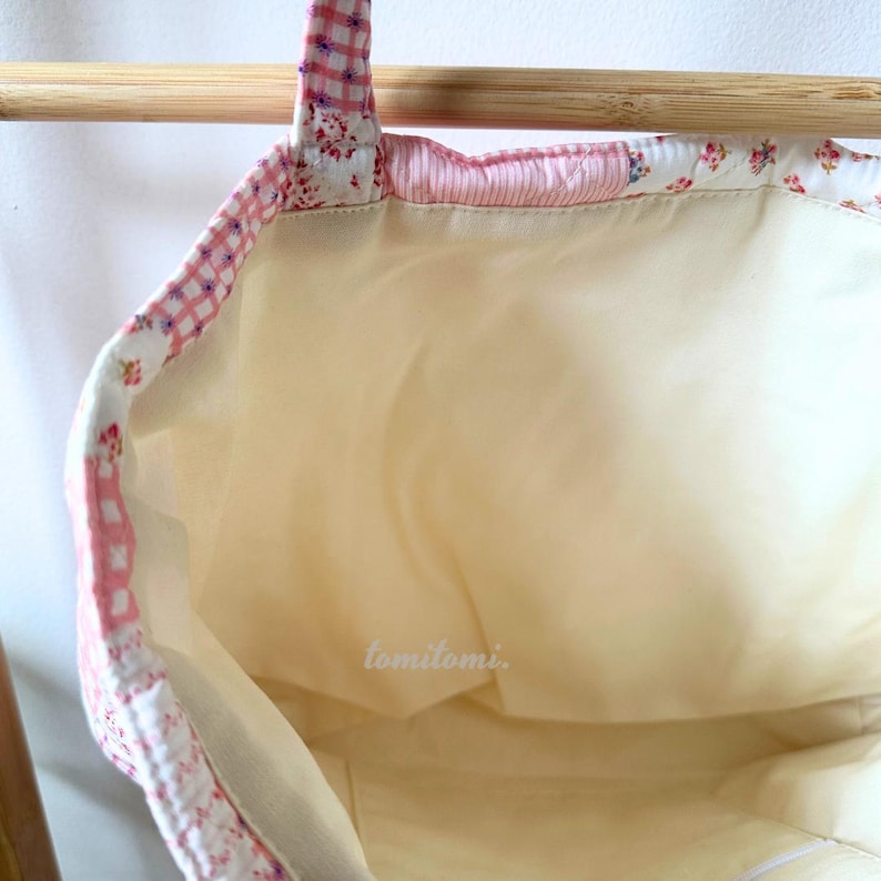 May include: A light yellow tote bag with a floral and gingham pattern trim. The bag's interior is a light yellow color, and the handle is made of the same patterned fabric. The text "tomitomi." is visible on the interior of the bag.