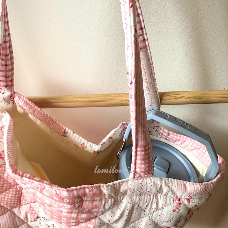May include: A quilted tote bag with pink and white floral and checkered patterns. The bag has a light beige interior and a gray container inside. The word "tomiton" is printed on the bag.