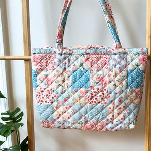 May include: Quilted tote bag with floral and polka dot patterns in pink, blue, and cream. The bag features a diamond-stitched design and matching handles. A stylish and practical accessory for carrying essentials.