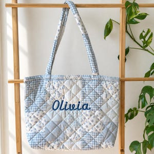 Op de afbeelding: Een gewatteerde tote bag met een patchworkontwerp van lichtblauw en wit gingham en bloemenpatronen. De tas heeft twee schouderbanden en de naam Olivia is in donkerblauw cursief schrift geborduurd op het voorpaneel in het midden.