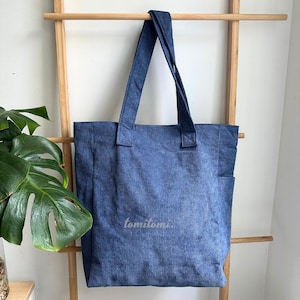 May include: A blue corduroy tote bag with two shoulder straps and a side pocket. The fabric features a vertical ribbed texture. The word "tomitomi." is printed in a small, light-colored font on the lower center of the front panel of the bag.