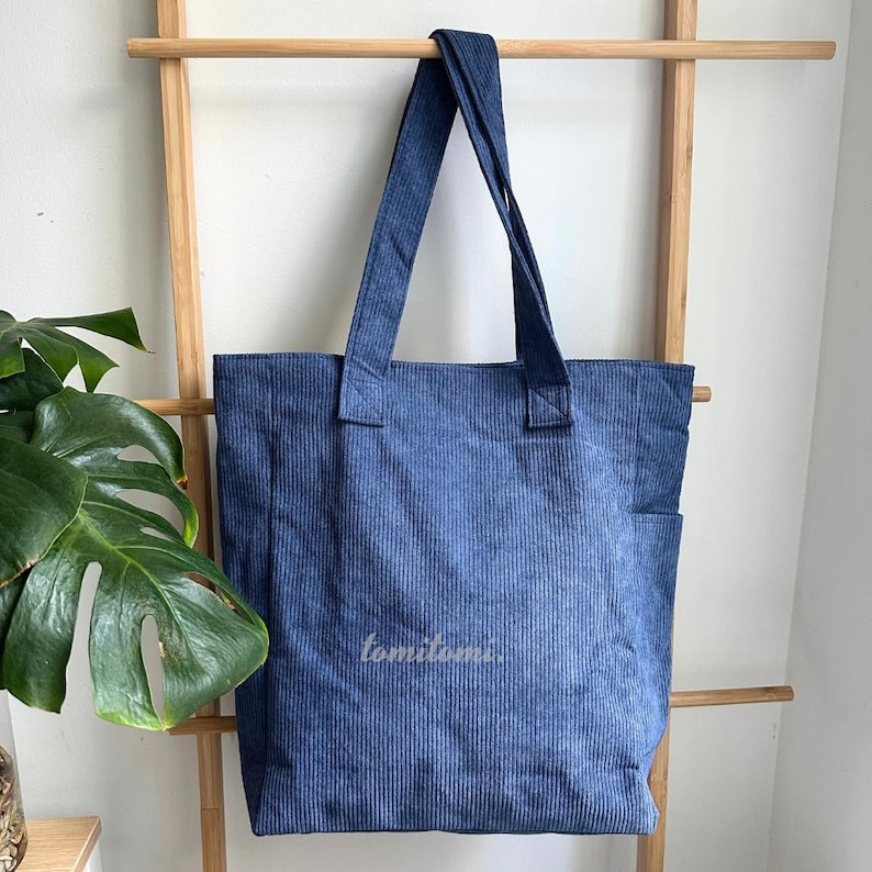 May include: A blue corduroy tote bag with two shoulder straps and a side pocket. The fabric features a vertical ribbed texture. The word "tomitomi." is printed in a small, light-colored font on the lower center of the front panel of the bag.