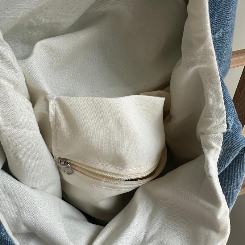May include: A denim tote bag with a cream-colored interior and a zippered pocket. The bag's exterior is a light blue denim, and the interior is a soft, light fabric. The zippered pocket has a silver zipper.