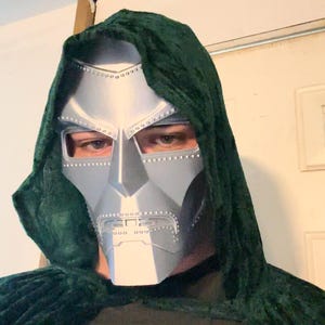 Può includere: Una maschera color argento con un design dettagliato, con aperture per gli occhi e un'area della bocca con ritagli rettangolari. La maschera fa parte di un costume, indossata con un mantello con cappuccio verde scuro.