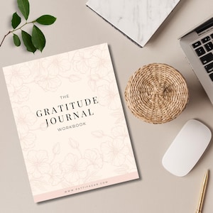 May include: A gratitude journal workbook with a floral design on a light peach background. The title "The Gratitude Journal Workbook" is in black text. A woven basket, laptop, mouse, and pen are also visible.