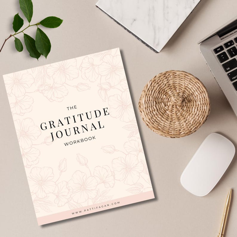 May include: A gratitude journal workbook with a floral design on a light peach background. The title "The Gratitude Journal Workbook" is in black text. A woven basket, laptop, mouse, and pen are also visible.