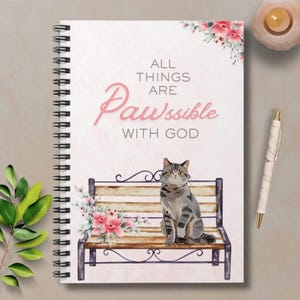 May include: Spiral-bound notebook with a cat design. The cover displays a grey tabby cat on a bench, floral decorations, and the text "ALL THINGS ARE PAWSSIBLE WITH GOD" in pink. A pen is placed next to the notebook.