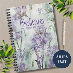 May include: Spiral-bound notebook with a floral iris design in shades of purple and green. The cover features the words "Believe and things will change" in a handwritten font. A pen with a marble design rests beside the notebook.