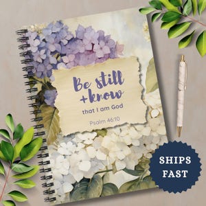 May include: Spiral-bound journal with a floral hydrangea design in shades of purple and white. The cover features the text "Be still + know that I am God" from Psalm 46:10. A marble-patterned pen rests beside the journal.