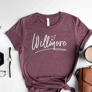 May include: A maroon t-shirt with the text "Willimore" in white script with a heart and arrow above the word. The shirt is laid flat on a white surface with a brown leather purse, a cup of coffee, a watch, a pair of glasses, and a small plant.