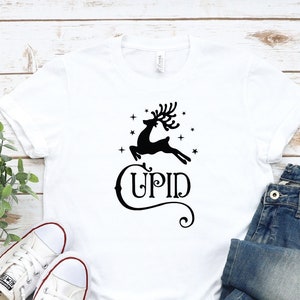 May include: White t-shirt with a black reindeer graphic and the word "CUPID" printed on it. The shirt is laid out on a white wooden surface with a pair of white sneakers and blue jeans.