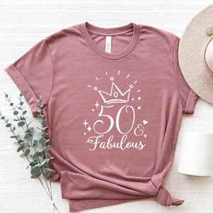 以下が含まれることがあります： 王冠、数字の50、そして「& Fabulous」という文字が白で描かれた、薄紫色のTシャツです。