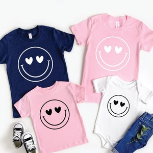 May include: A set of four t-shirts featuring a smiley face with hearts for eyes. The shirts are in navy blue, pink, and white.