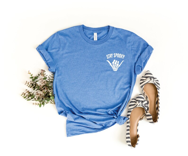 May include: A blue t-shirt with the text "Stay Spooky" and a skeleton hand making a shaka sign. The shirt is laid flat on a white background with a sprig of greenery and two black and white striped flats.