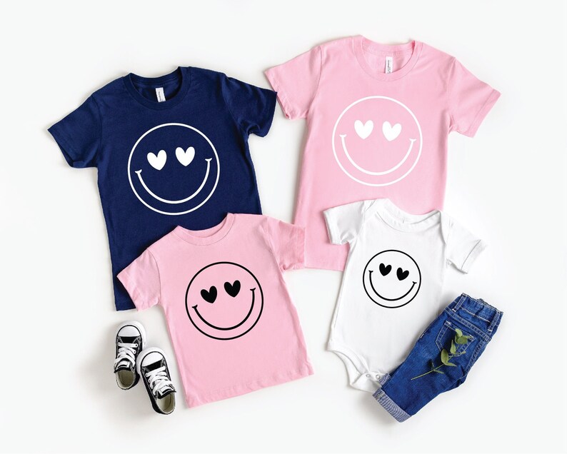 May include: A set of four t-shirts featuring a smiley face with hearts for eyes. The shirts are in navy blue, pink, and white.