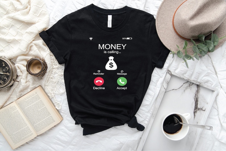 K&ouml;nnte beinhalten: Schwarzes T-Shirt mit einer wei&szlig;en Grafik eines Telefonbildschirms. Der Bildschirm zeigt "MONEY is calling..." und ein Geldbeutelsymbol mit einem Dollarzeichen. Der Telefonbildschirm hat zwei Schaltfl&auml;chen: "Decline" und "Accept".