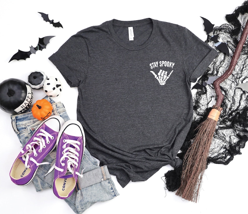 May include: A dark gray t-shirt with the text 'Stay Spooky' and a skeleton hand making a shaka sign. The t-shirt is laid flat on a white surface. There are purple converse shoes, a broom, and Halloween decorations in the background.