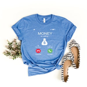 K&ouml;nnte beinhalten: Ein blaues T-Shirt mit einer wei&szlig;en Grafik eines Telefonbildschirms, auf dem "MONEY is calling--" steht, mit einem Geldbeutelsymbol und zwei Kn&ouml;pfen, auf denen "Decline" und "Accept" steht.