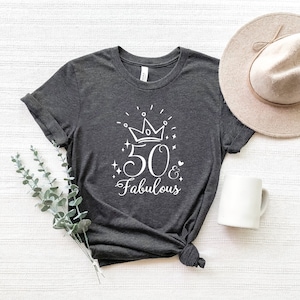 以下が含まれることがあります： 暗いグレーのTシャツに、白いグラフィックデザインで「50 & Fabulous」と書いてあり、数字の50の上に王冠があります。