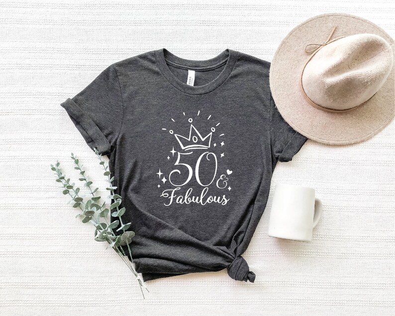 以下が含まれることがあります： 暗いグレーのTシャツに、白いグラフィックデザインで「50 & Fabulous」と書いてあり、数字の50の上に王冠があります。