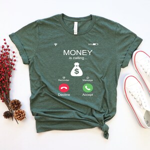K&ouml;nnte beinhalten: Ein gr&uuml;nes T-Shirt mit einer wei&szlig;en Grafik eines Telefonbildschirms, auf dem "MONEY is calling..." steht, mit einem Geldbeutelsymbol und Kn&ouml;pfen mit der Aufschrift "Decline" und "Accept".
