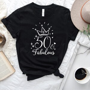 以下が含まれることがあります： 白い王冠、数字の50、そして「& Fabulous」という文字が書かれた白いグラフィックデザインが施された黒いTシャツ。