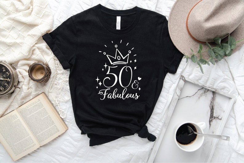 以下が含まれることがあります： 白い王冠、数字の50、そして「& Fabulous」という文字が書かれた白いグラフィックデザインが施された黒いTシャツ。