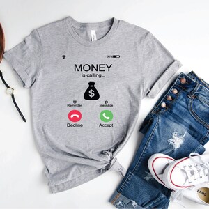 K&ouml;nnte beinhalten: Graues T-Shirt mit einem Grafikdruck eines Telefonbildschirms, auf dem "MONEY is calling..." steht, mit einem Geldbeutelsymbol und Kn&ouml;pfen mit der Aufschrift "Decline" und "Accept".
