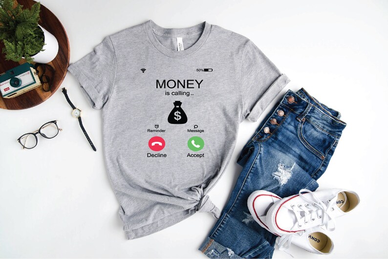 K&ouml;nnte beinhalten: Graues T-Shirt mit einem Grafikdruck eines Telefonbildschirms, auf dem "MONEY is calling..." steht, mit einem Geldbeutelsymbol und Kn&ouml;pfen mit der Aufschrift "Decline" und "Accept".