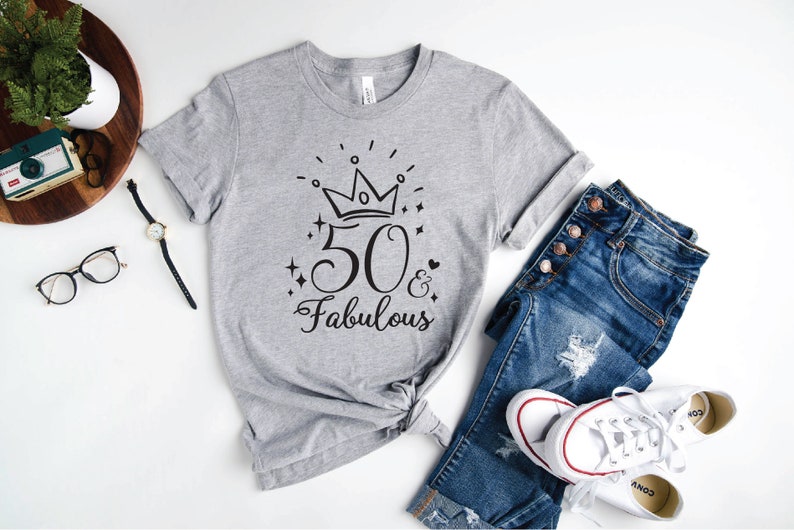 以下が含まれることがあります： 黒い王冠と「50 & Fabulous」の文字がプリントされたグレーのTシャツ。Tシャツは前で結ばれています。