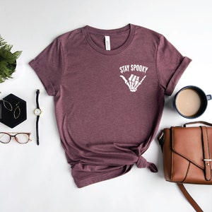 May include: A maroon t-shirt with the text "Stay Spooky" and a white skeleton hand making a shaka sign. The shirt is laid out on a white surface with a watch, glasses, earrings, a plant, a brown purse, and a cup of coffee.