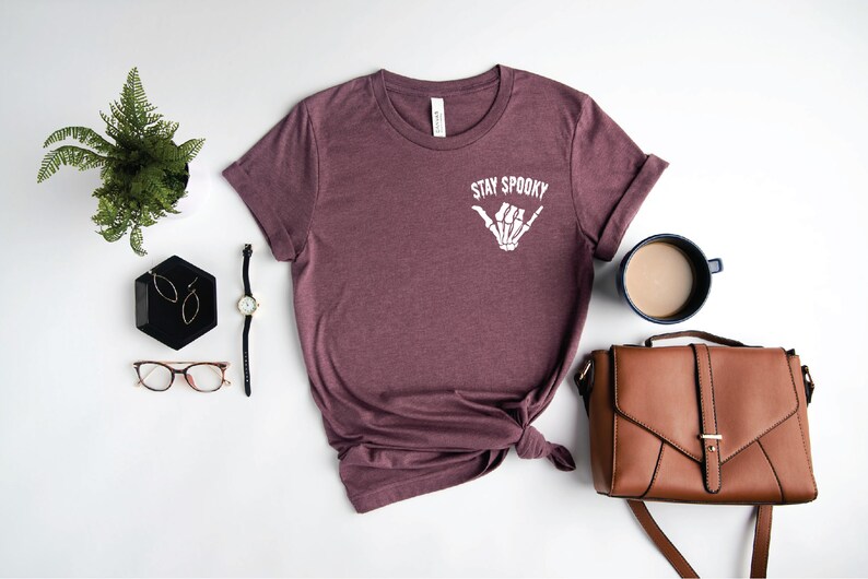 May include: A maroon t-shirt with the text "Stay Spooky" and a white skeleton hand making a shaka sign. The shirt is laid out on a white surface with a watch, glasses, earrings, a plant, a brown purse, and a cup of coffee.