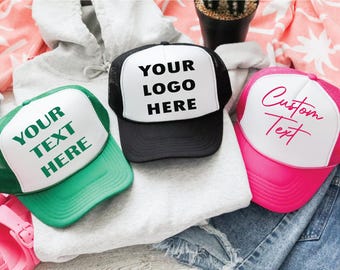 May include: Three trucker hats with white front panels and mesh backs. One hat is green with "YOUR TEXT HERE" printed in green. Another is black with "YOUR LOGO HERE" in black. The third is pink with "Custom Text" in pink.