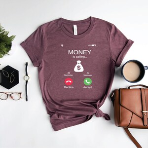 K&ouml;nnte beinhalten: Ein maronfarbenes T-Shirt mit einer wei&szlig;en Grafik eines Telefonbildschirms, auf dem "MONEY is calling..." steht, mit einem Geldbeutelsymbol und zwei Kn&ouml;pfen mit der Aufschrift "Decline" und "Accept".