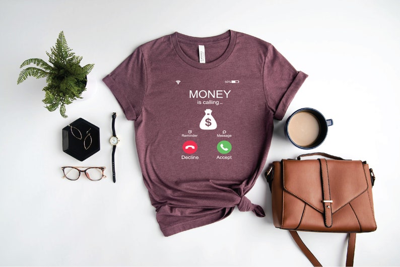 K&ouml;nnte beinhalten: Ein maronfarbenes T-Shirt mit einer wei&szlig;en Grafik eines Telefonbildschirms, auf dem "MONEY is calling..." steht, mit einem Geldbeutelsymbol und zwei Kn&ouml;pfen mit der Aufschrift "Decline" und "Accept".