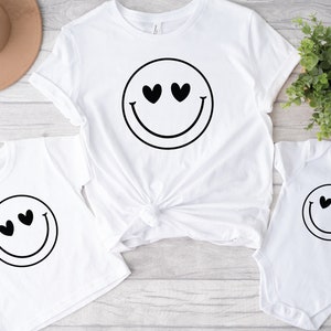 May include: Three white t-shirts featuring a black smiley face design. The smiley face has hearts for eyes. The shirts are in different sizes.