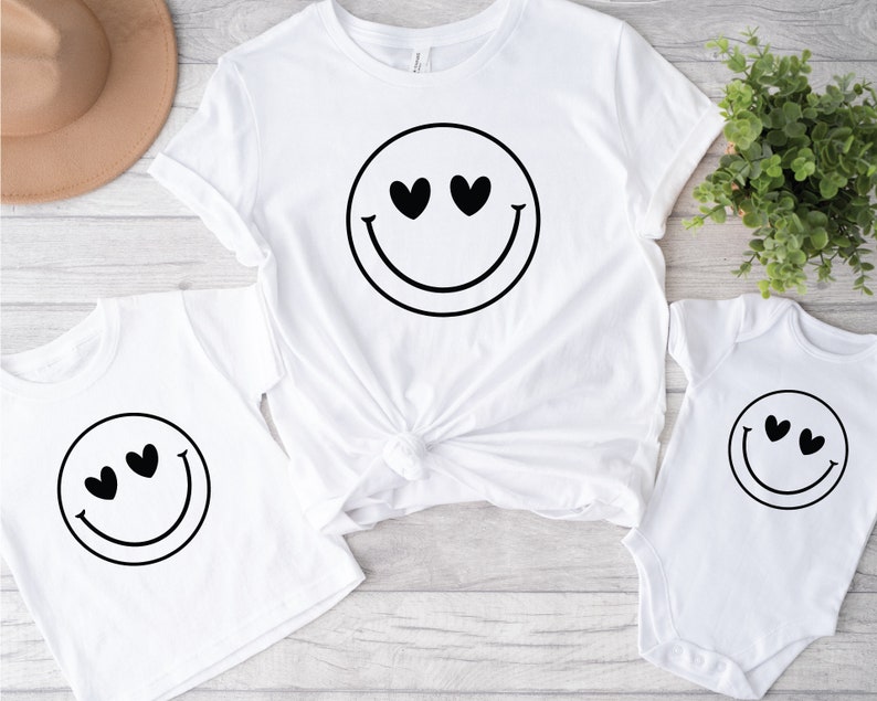 May include: Three white t-shirts featuring a black smiley face design. The smiley face has hearts for eyes. The shirts are in different sizes.