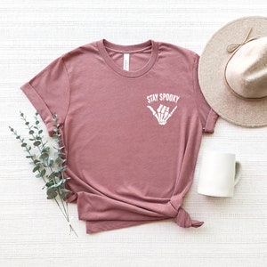 May include: A mauve t-shirt with a white skeleton hand making a shaka sign and the text "Stay Spooky".
