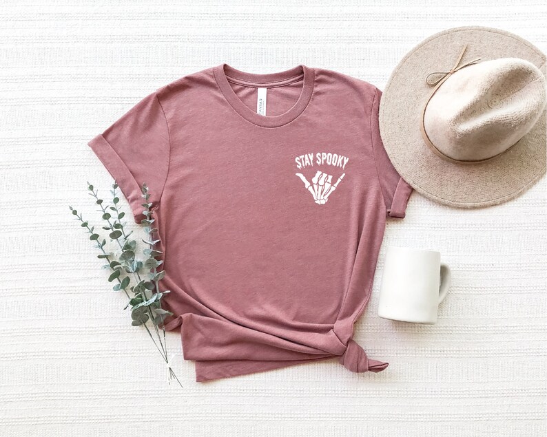 May include: A mauve t-shirt with a white skeleton hand making a shaka sign and the text "Stay Spooky".