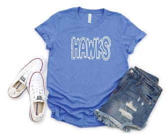 May include: A royal blue t-shirt with white text that reads "HAWKS" in a stylized font. The shirt is shown with a pair of white sneakers and a pair of denim shorts.