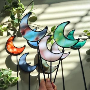 May include: A collection of colorful stained glass crescent moon garden stakes. The moons feature shades of orange, blue, green, and pink, with black metal stems. The glass has a textured appearance, and the stakes are arranged in a decorative display.