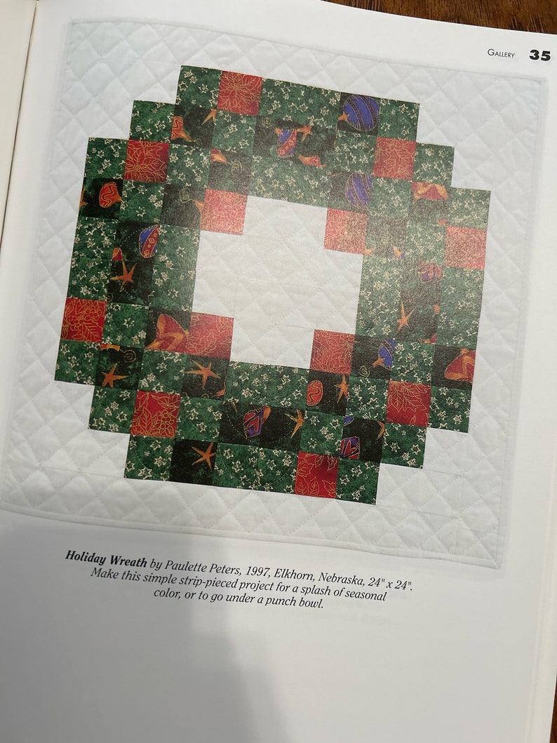 K&ouml;nnte beinhalten: Ein gr&uuml;ner, roter und blauer Patchwork-Decken in Form eines Kranzes. Die Decke besteht aus Quadraten und ist 61 cm x 61 cm gro&szlig;. Der Text "Holiday Wreath by Paulette Peters, 1997, Elkhorn, Nebraska, 24" x 24". Make this simple strip-pieced project for a splash of seasonal color, or to go under a punch bowl." ist unter der Decke gedruckt.