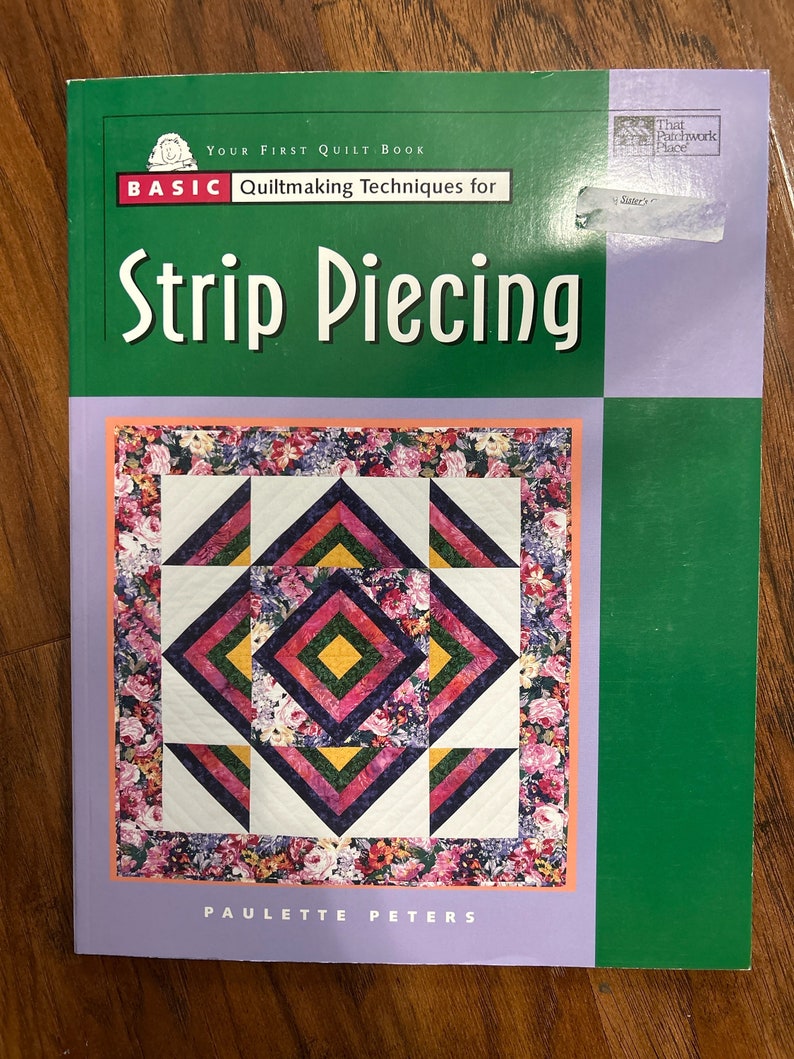 K&ouml;nnte beinhalten: Ein gr&uuml;nes und violettes Buch mit dem Titel "Your First Quilt Book: Basic Quiltmaking Techniques for Strip Piecing" von Paulette Peters. Der Umschlag zeigt eine farbenfrohe Steppdecke mit einem Blumenmuster.