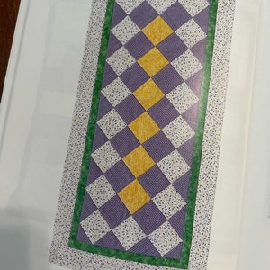 K&ouml;nnte beinhalten: Ein lila und gelber Tischl&auml;ufer mit einem wei&szlig;en Blumenmuster am Rand. Der L&auml;ufer besteht aus abwechselnden Quadraten aus lila und gelbem Stoff. Der Text "Table Runner by Paulette Peters, 1997. Elkhorn, Nebraska. 18" x 40". Trip Around the World, a traditional favourite that lends itself to a long, narrow version." ist unter dem L&auml;ufer gedruckt.