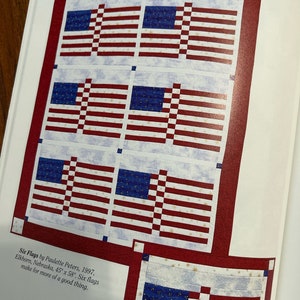 K&ouml;nnte beinhalten: Sechs amerikanische Flaggen sind in einem Quiltmuster zusammengen&auml;ht. Die Flaggen sind rot, wei&szlig; und blau mit wei&szlig;em Hintergrund und goldenen Sternen. Der Quilt ist 45 Zoll mal 58 Zoll gro&szlig;. Der Titel "Six Flags" steht unter dem Quilt.