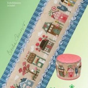 Könnte beinhalten: Ein Kreuzstichmuster mit einem "Blooming Tiny Town" Design. Das Muster zeigt ein langes, schmales, gesticktes Stück mit winzigen Häusern, Blumen und einem blauen Vogel. Ein kleines, rundes Nadelkissen mit passendem Design ist ebenfalls abgebildet.