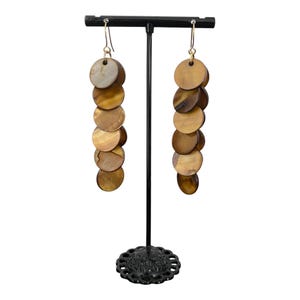 May include: A pair of long drop earrings featuring a vertical chain of six circular, iridescent brown shell discs. Each earring hangs from a thin gold-toned metal hook, suspended on a black T-shaped jewellery display stand with a decorative base.