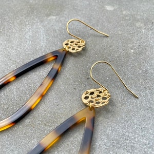 Może przedstawiać: Para złotych kolczyków w kolorze żółwim. Kolczyki mają kształt łzy i są ozdobione złotym wzorem filigranowym u góry.