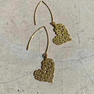 Könnte beinhalten: Ein Paar goldene Filigran-Herzohrringe mit zarten Drahtschlaufen. Die Ohrringe hängen an einer hellgrauen Oberfläche.